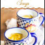 A cup of pumpkin soup topped with foamed Half & Half, sitting on a placemat with a spoon.