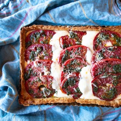 This Tomato Caprese Tart is easy to make with fresh peak season tomatoes, mozzarella cheese, and a pesto - all tucked into a tart shell. Use it for an appetizer, side dish or vegetarian dinner.