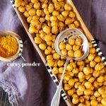 A spoonful of crispy roasted chickpeas that have been dusted with curry powder. The spoon rests on a tray of more chickpeas, and a tiny wooden bowl of curry powder.