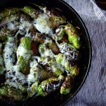 A cast iron skillet of blistered shishito peppers topped with melted cheese.