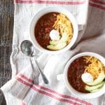 Two bowls of vegan chili con carne topped with avocado slices, vegan sour cream and vegan cheddar cheese.