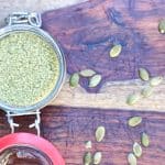 A jar of ground up pumpkin seeds, with a few pumpkin seeds scattered around it.
