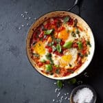 A skillet of poached eggs in a spicy tomato sauce and black beans, topped with oregano and basil.