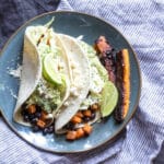 A plate of Cuban seasoned tacos filled with sweet potatoes, black beans, shredded cabbage and avocado sauce.