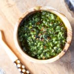 A wooden bowl of cilantro chimichurri with red pepper flakes on a cutting board.