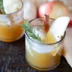 Two glasses of apple cider bourbon cocktail garnished with rosemary, cinnamon sticks, and apple slices.