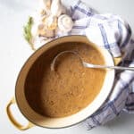 A big pot of mushroom gravy with a few mushrooms and a sprig of rosemary beside it.